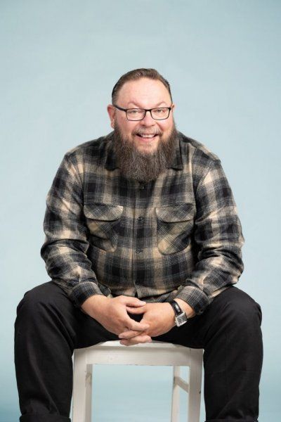 Hymyilevä parrakas ja silmälasit omaava henkilö istuu tuolilla ruudullinen flanellipaita yllään vaaleansinistä taustaa vasten.