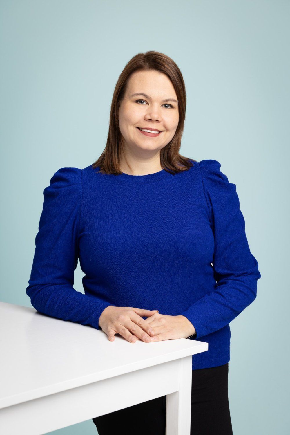 Kirkkaansiniseen pitkähihaiseen paitaan pukeutunut henkilö nojaa valkoiseen pöytään vaaleansinistä taustaa vasten.