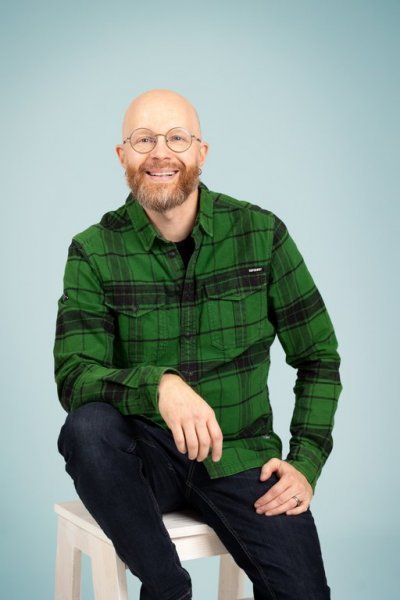Hymyilevä, silmälasit ja parrakas henkilö istuu jakkaralla vaaleansinistä taustaa vasten yllään vihreä ruutupaita.