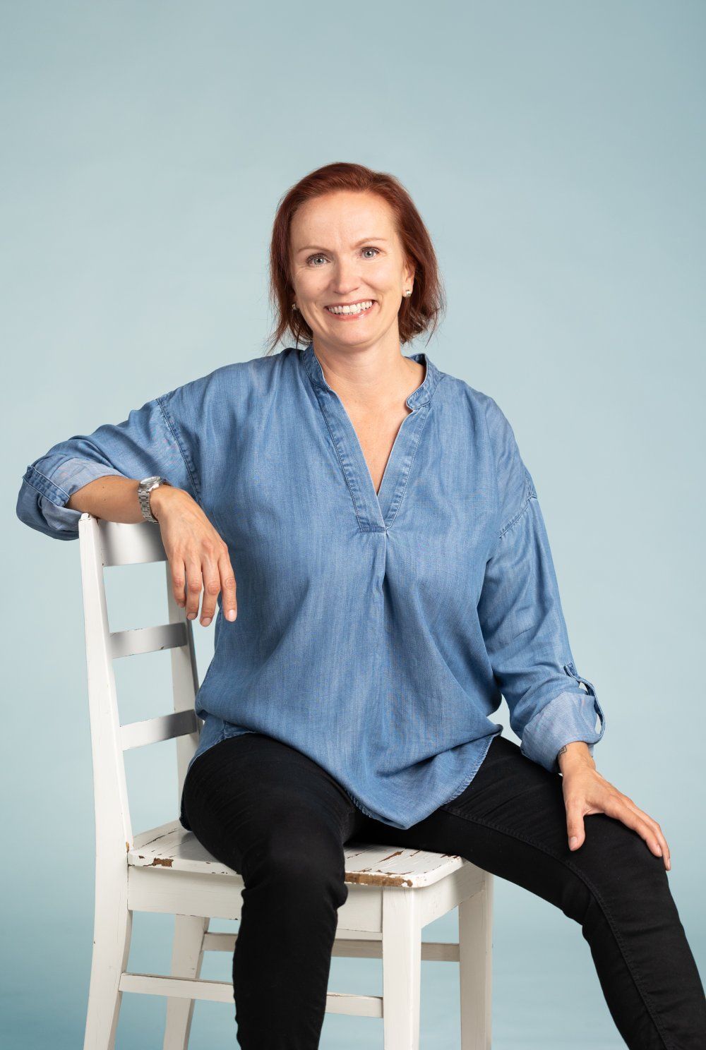 Punatukkainen henkilö, jolla on sininen farkkupaita ja mustat housut, istuu valkoisella tuolilla sinistä taustaa vasten.