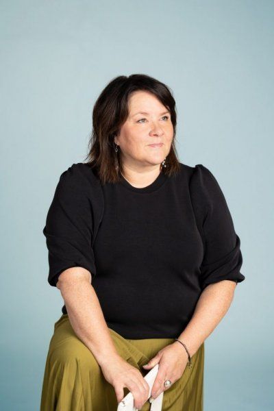 Hartioille ulottuvat tummat hiukset, yllään musta paita ja vihreä hame, katsoo sivuun sinistä seinää vasten.