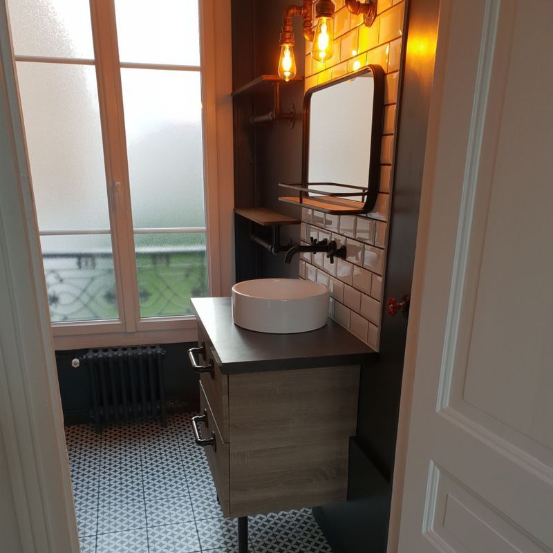 Salle de bains avec meuble-lavabo, miroir, éclairage industriel et mur carrelé. Une fenêtre en verre dépoli.