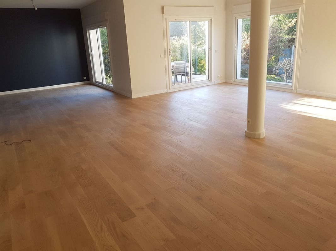 Pièce vide avec parquet, mur sombre, grandes fenêtres et colonne blanche.