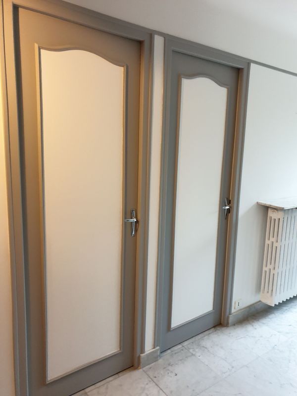 Deux portes blanches avec des encadrements et des poignées grises, et un radiateur sur un sol carrelé.