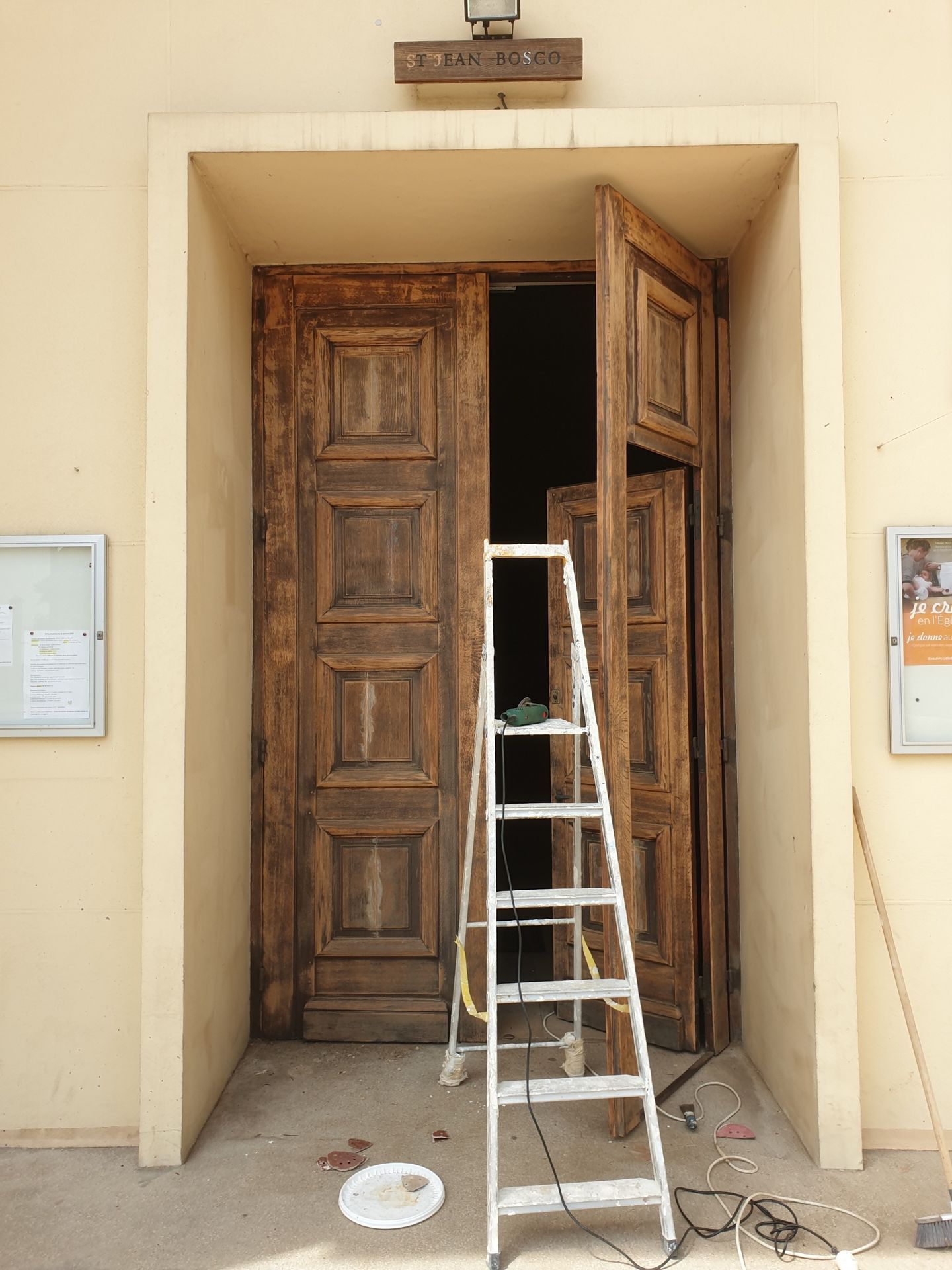 Les portes en bois de l'église sont entrouvertes, laissant entrevoir un intérieur sombre. Une échelle est appuyée contre la porte, des outils sont éparpillés au sol.