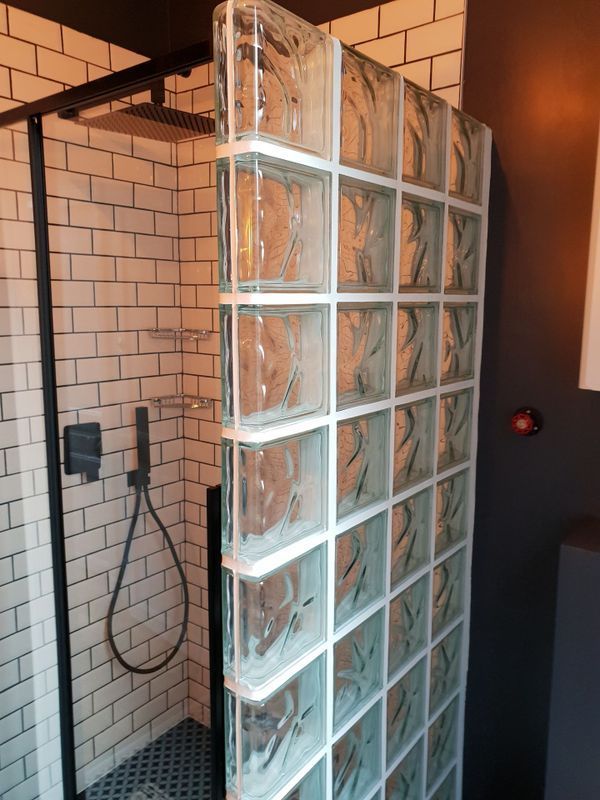 Douche avec paroi en pavés de verre, murs carrelés blancs, robinetterie noire et pommeau de douche carré effet pluie.