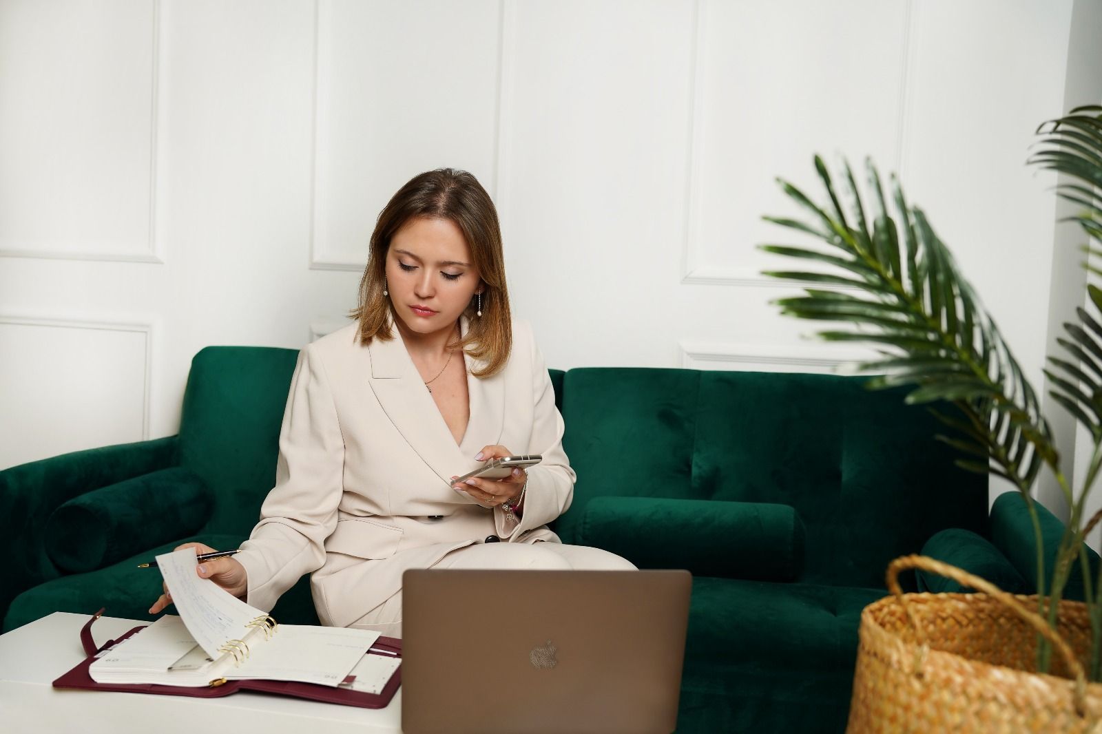 Una mujer con traje de color crema se sienta en un sofá verde, trabajando en una computadora portátil y un cuaderno.