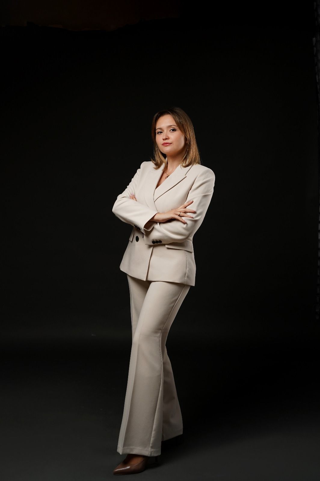 Mujer con traje pantalón beige, con los brazos cruzados, posa sobre un fondo oscuro.