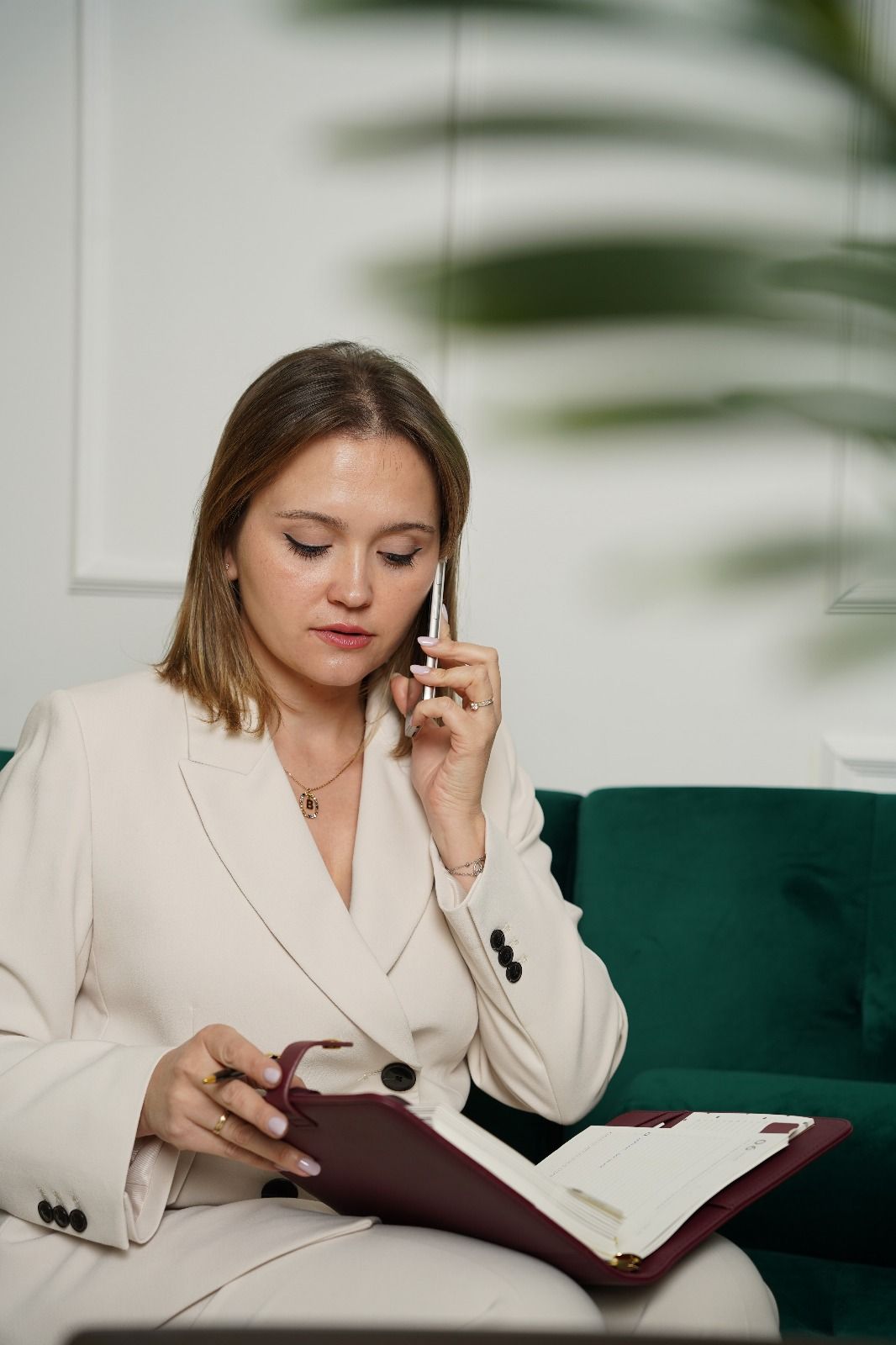 Mujer con traje blanco hablando por teléfono y mirando un cuaderno en un sofá verde.