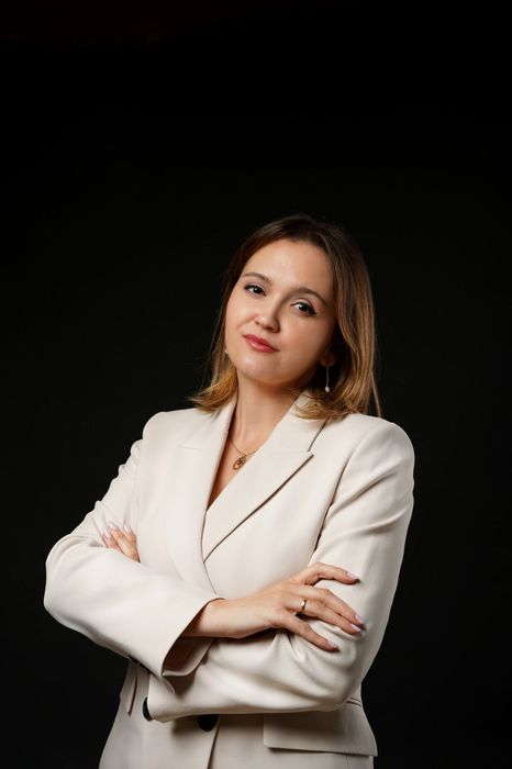 Mujer con un blazer color crema, con los brazos cruzados y sonriendo levemente sobre un fondo negro.