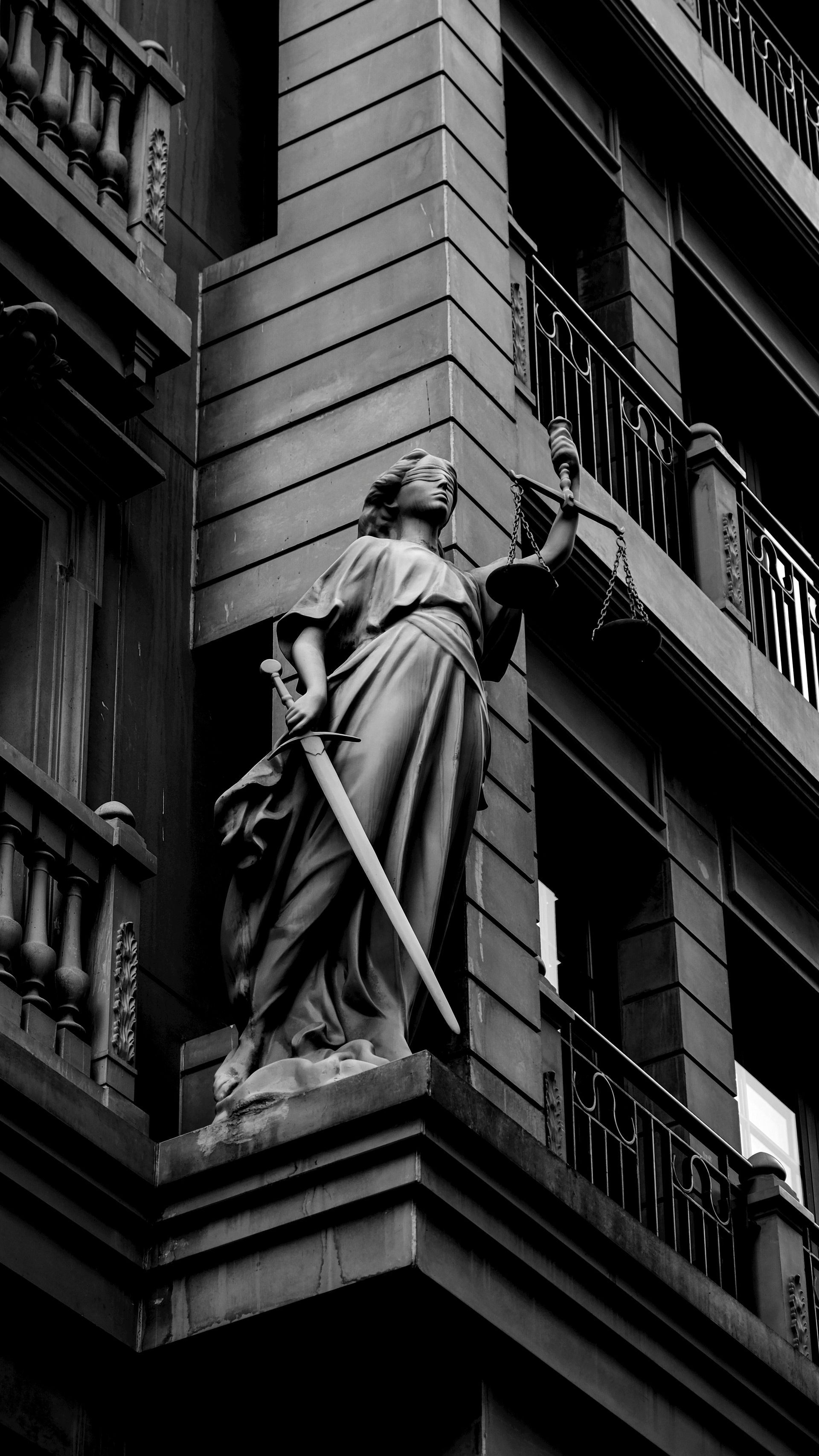 Estatua de la Dama de la Justicia en la esquina del edificio, sosteniendo espada y balanza, en blanco y negro.