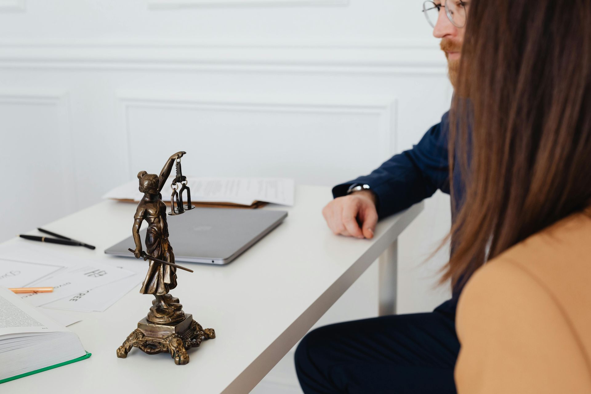 Estatua de Lady Justice sobre un escritorio con una computadora portátil; dos personas están sentadas cerca.