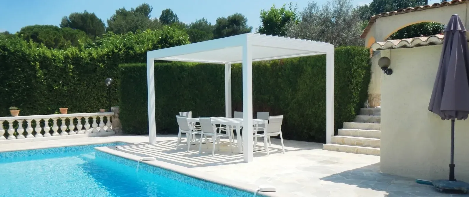 Une pergola blanche avec une table et des chaises se trouve près d'une piscine. On aperçoit une haie et un escalier en arrière-plan.