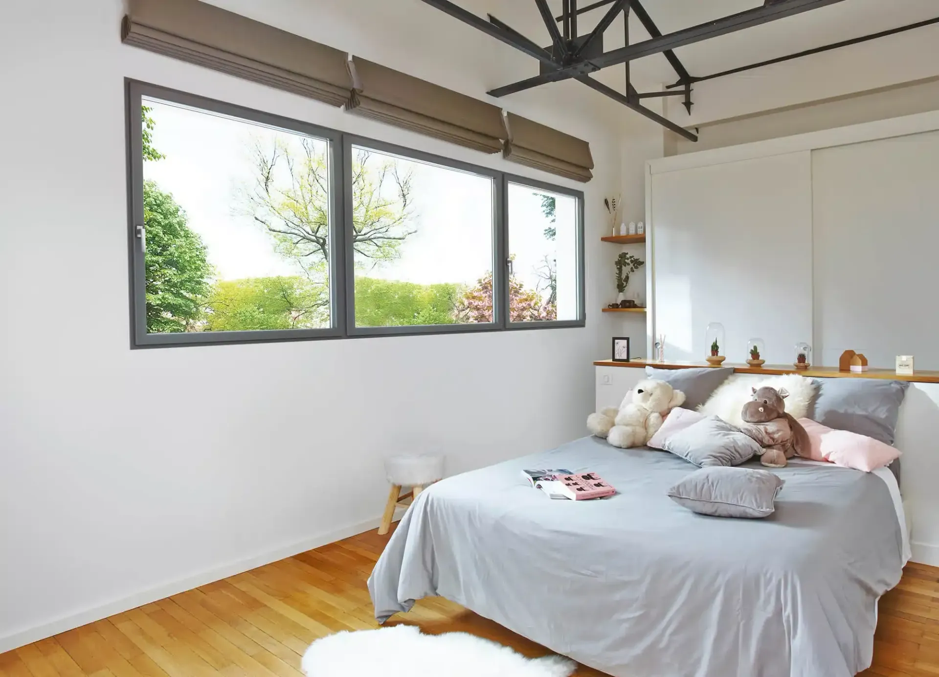 Chambre avec fenêtres à cadre gris, lit, parquet, murs blancs. Stores romains beiges et peluches visibles.