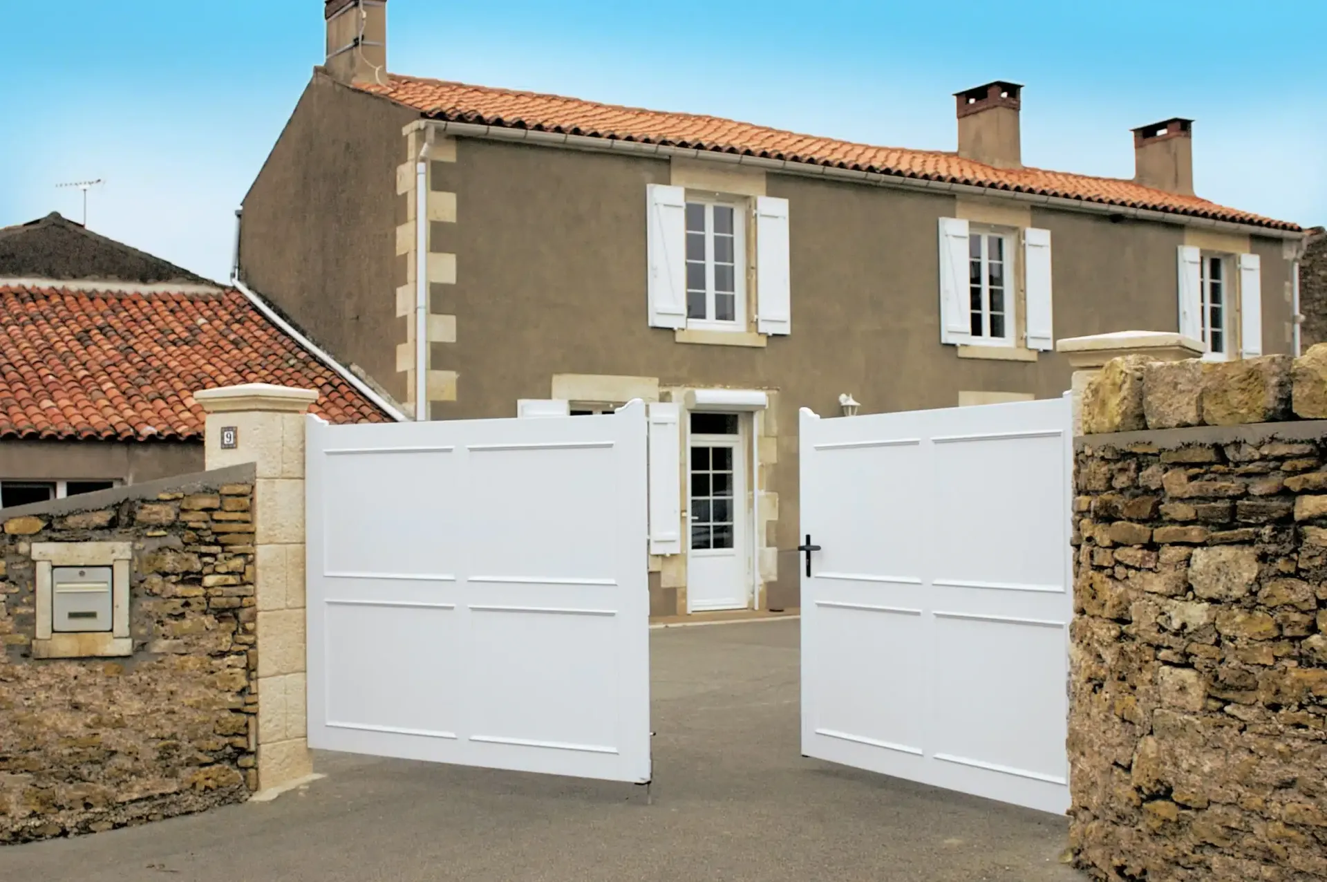 Un portail blanc s'ouvre sur une maison en pierre avec des volets, un toit en briques et une allée.