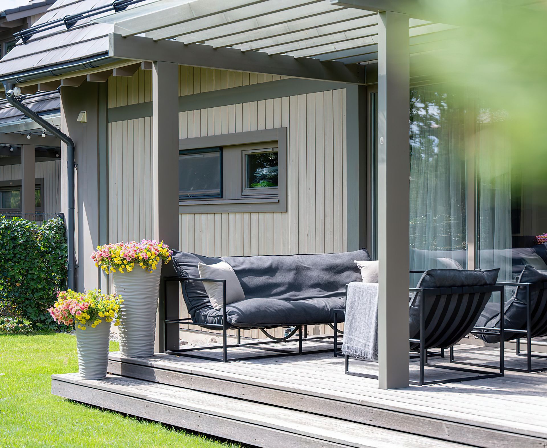 Patio avec mobilier gris, pergola et fleurs en pot. Terrasse en bois devant une maison.
