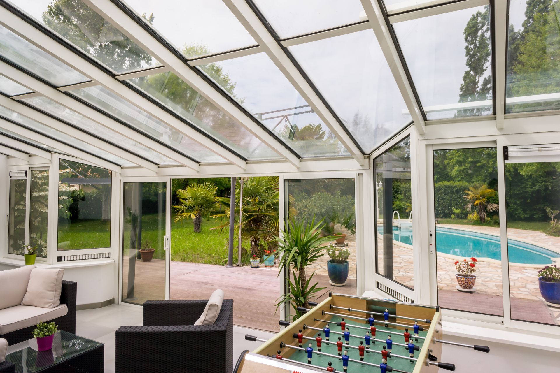 Véranda avec toit en verre donnant sur un jardin avec piscine. Le mobilier comprend un canapé, des fauteuils et un baby-foot.
