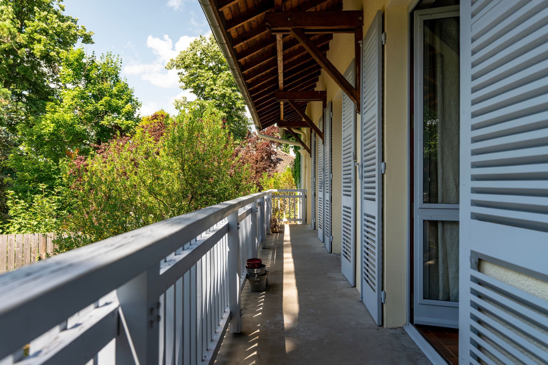 Balcon avec rambarde et volets blancs, arbres en arrière-plan, journée ensoleillée.