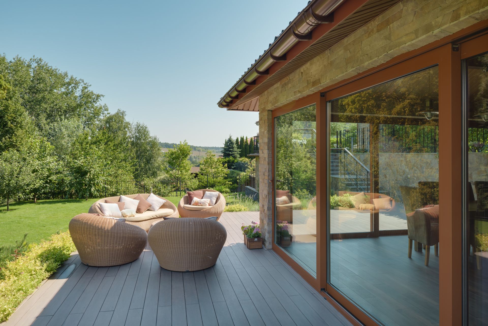 Terrasse en bois avec mobilier en osier donnant sur un jardin verdoyant et arboré, à côté d'un bâtiment aux grandes fenêtres.