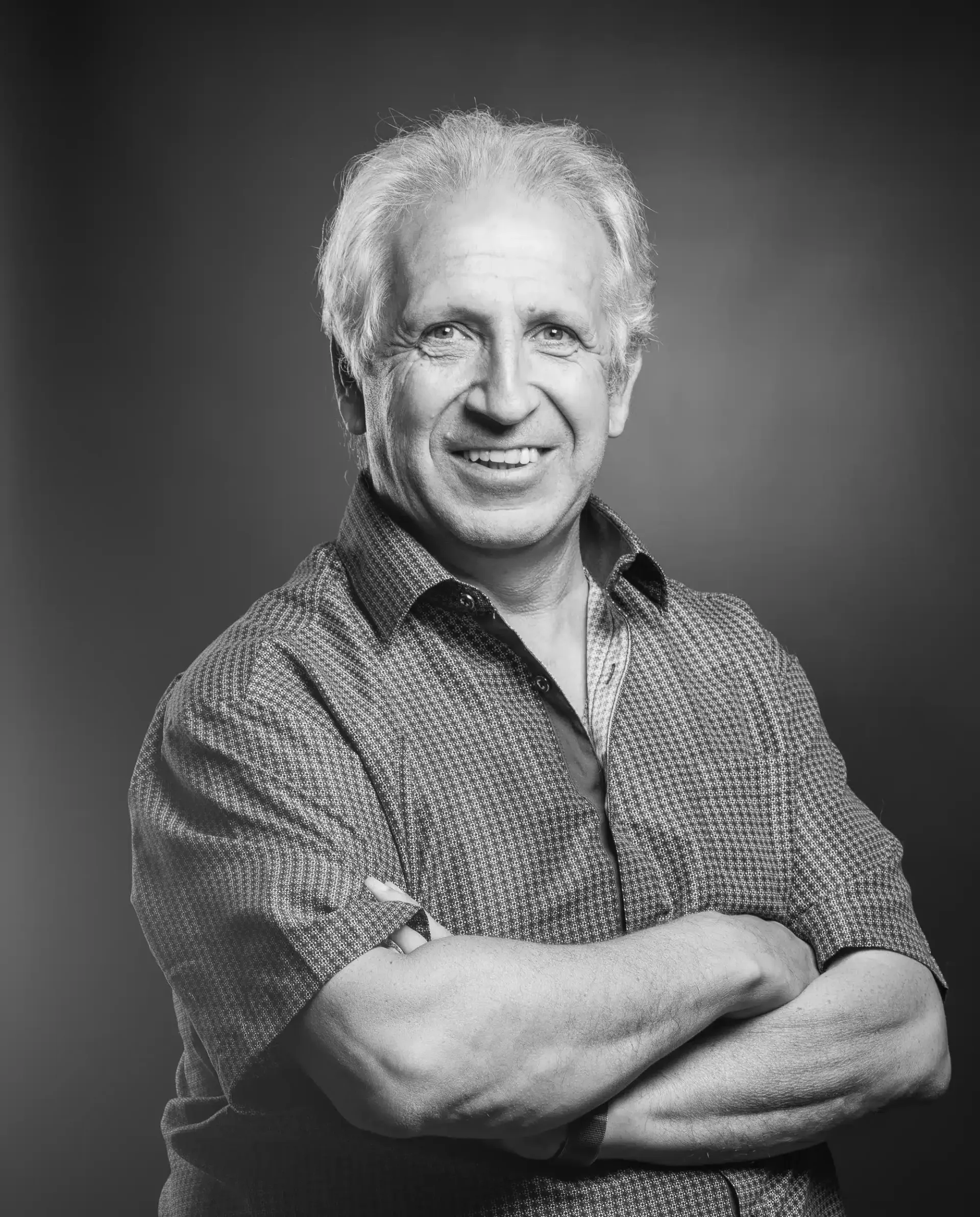 Un homme, les bras croisés, sourit devant un fond sombre et porte une chemise à motifs.