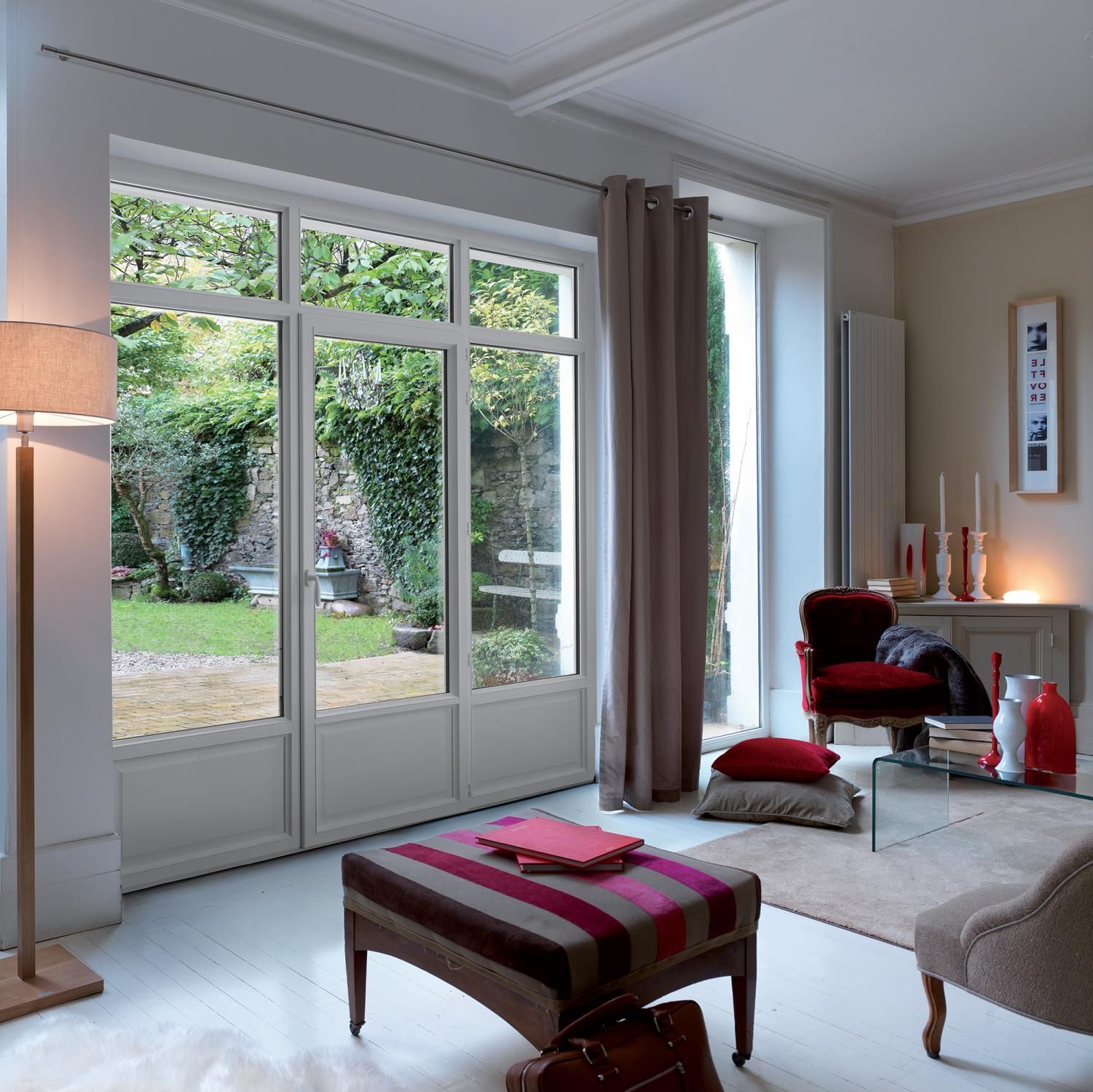 Salon avec de grandes fenêtres donnant sur un jardin. Il comprend un pouf à motifs, un fauteuil rouge et un lampadaire.