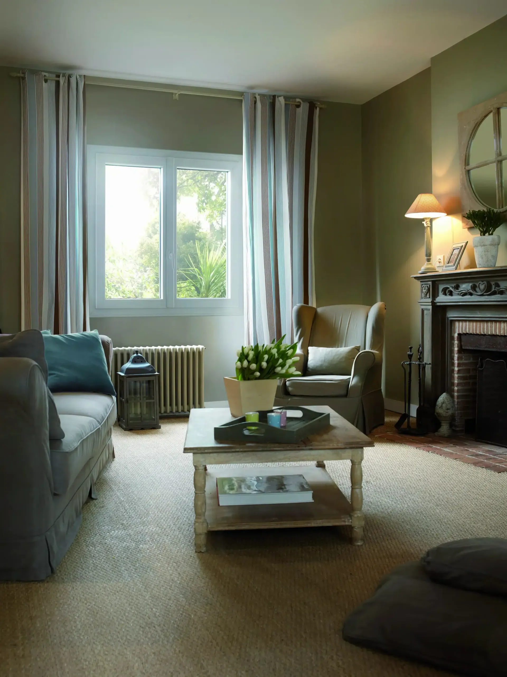 Salon aux tons neutres, canapé, fauteuil, cheminée et fenêtre avec rideaux à rayures.