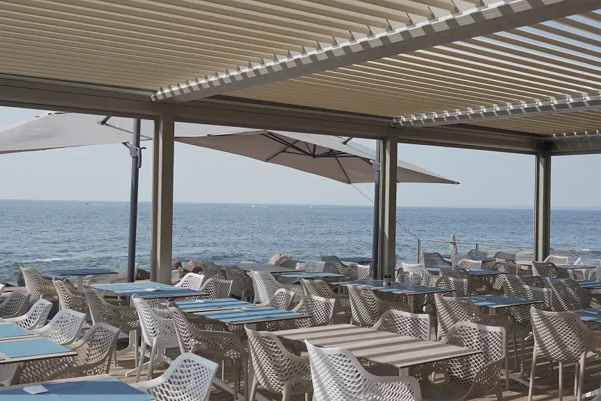 Restaurant en plein air avec tables et chaises sous une pergola à treillis donnant sur l'océan.