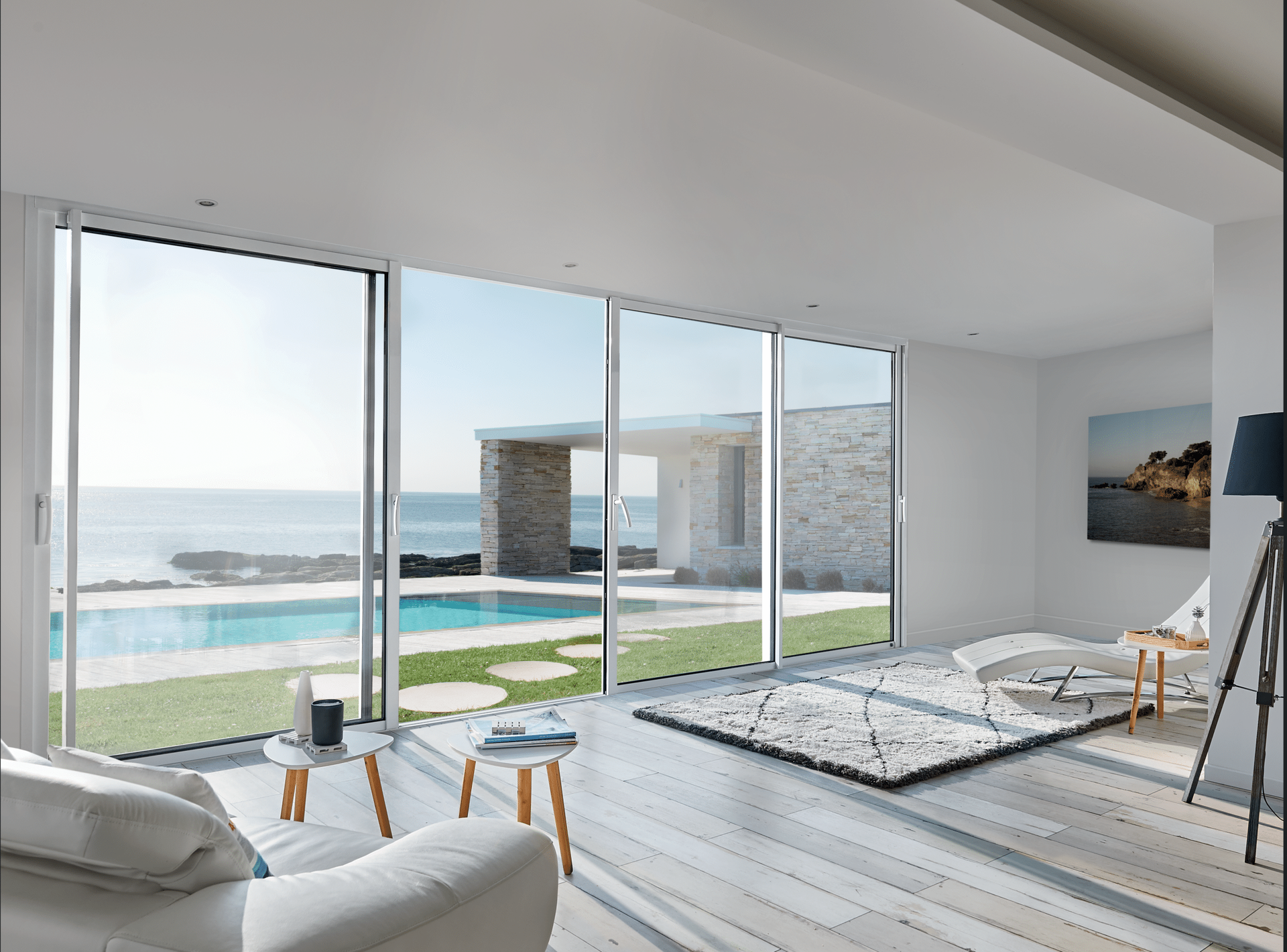 Salon avec de grandes fenêtres donnant sur une piscine, l'océan et un bâtiment en pierre. Murs blancs, parquet et mobilier en bois.