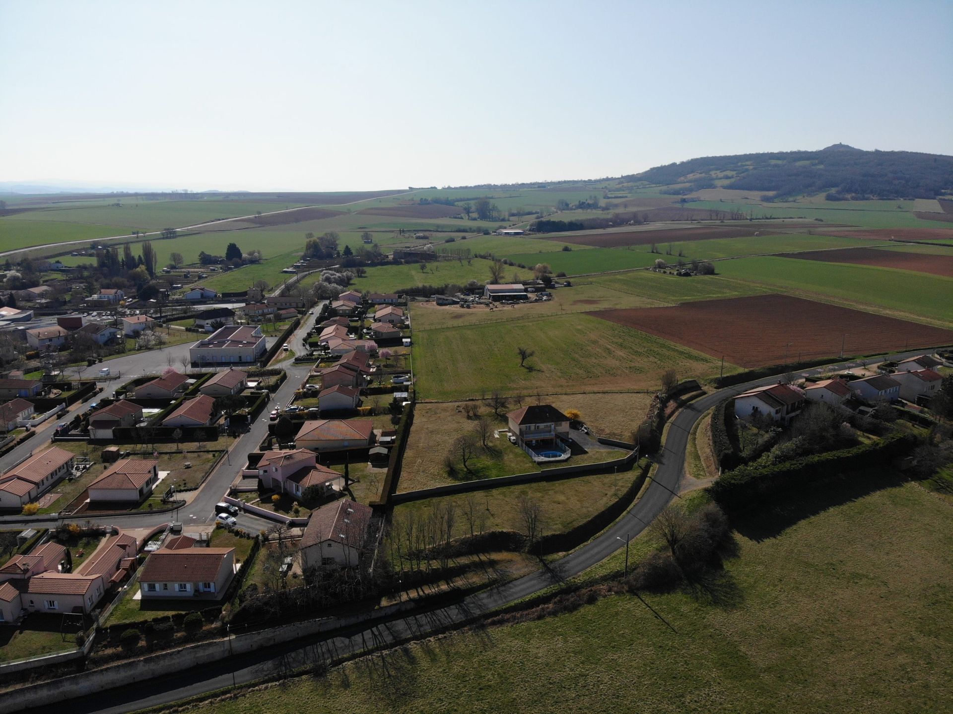 Vue aérienne d'un village avec des maisons, des routes et des champs agricoles sous un ciel ensoleillé.