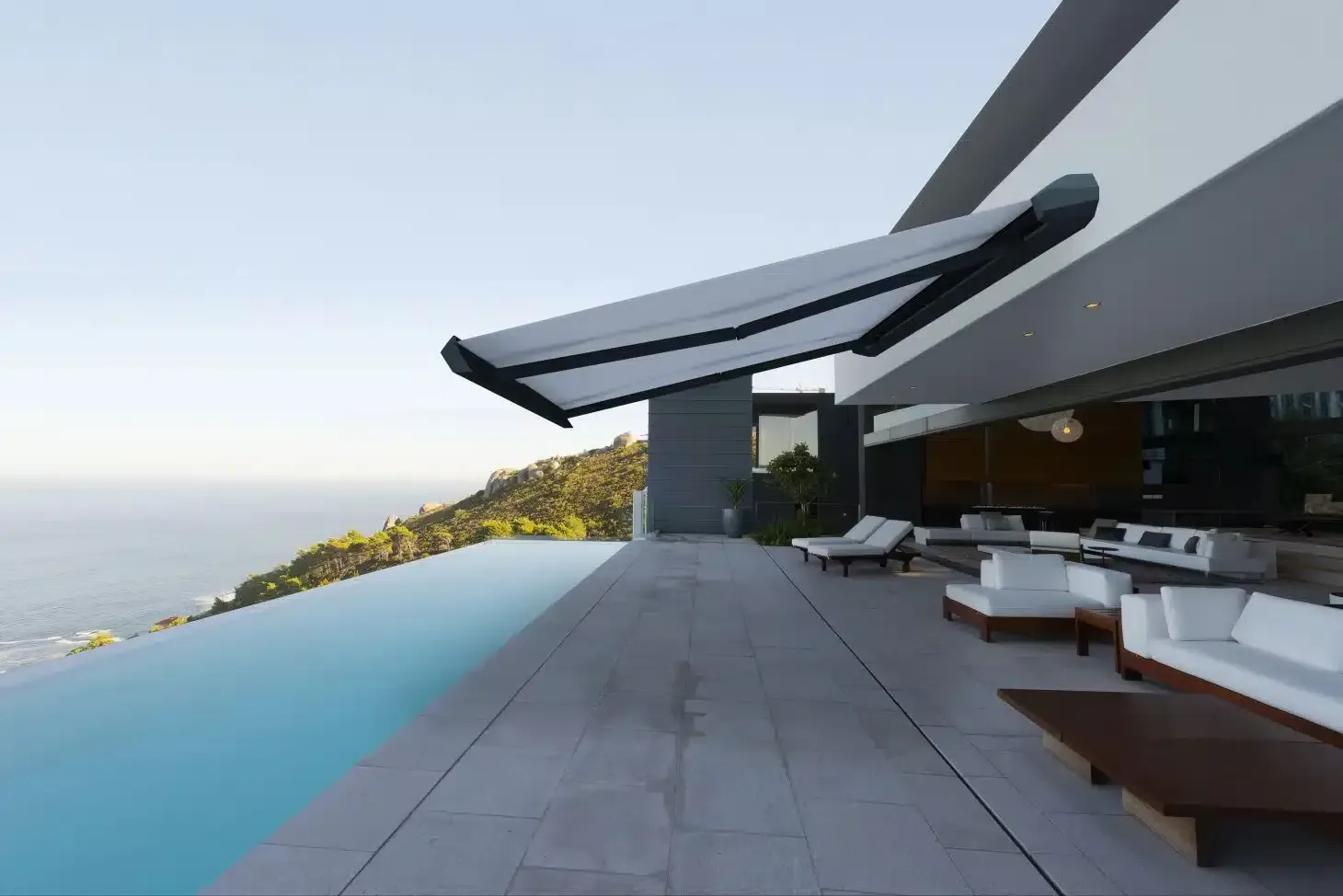 Maison moderne avec un store rétractable au-dessus d'une terrasse extérieure donnant sur une piscine à débordement et l'océan.
