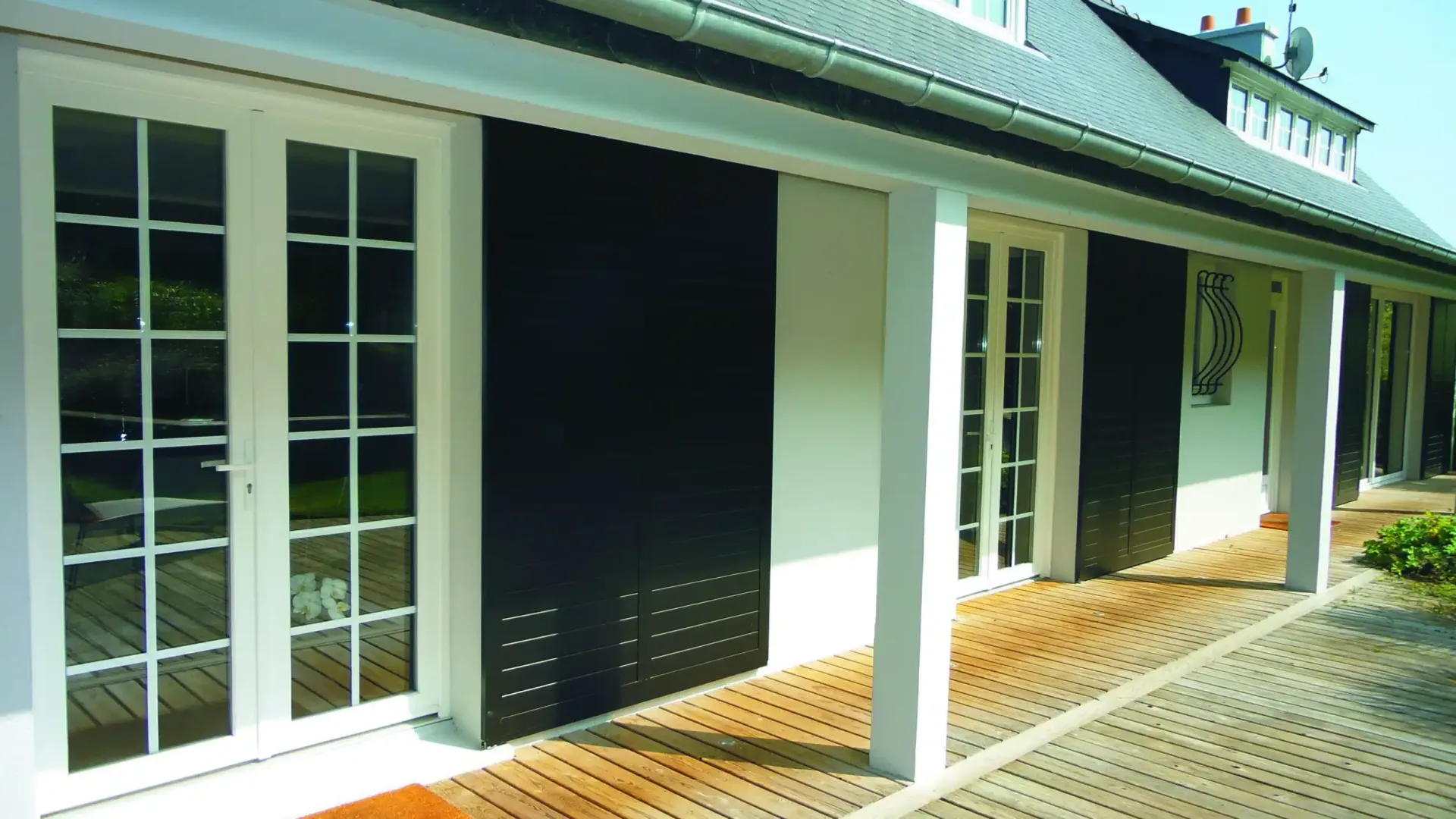 Maison blanche avec volets noirs sur portes vitrées, donnant sur une terrasse en briques.