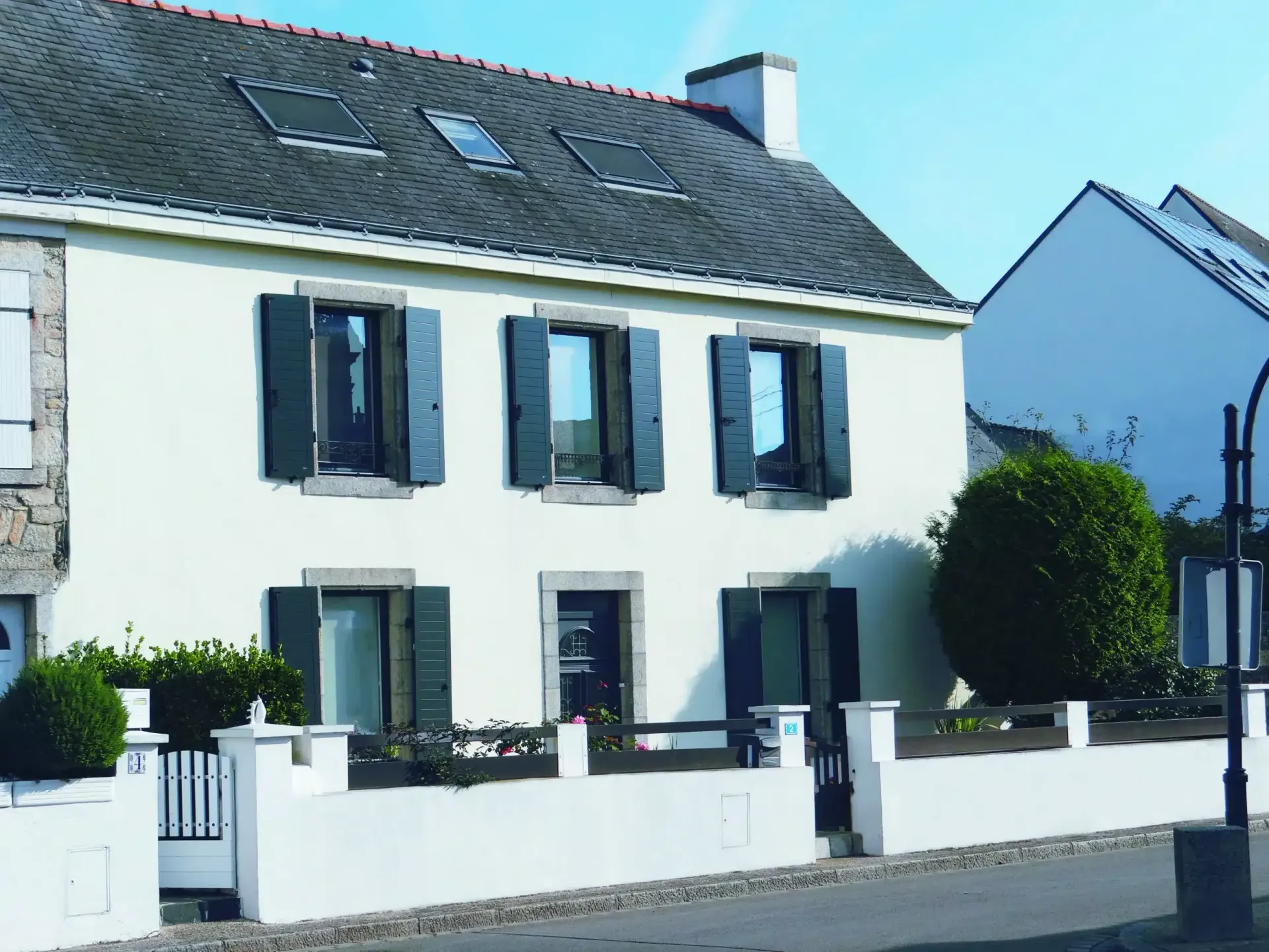 Maison blanche à deux étages avec des volets bleus, un toit gris et une petite cour derrière un mur blanc.