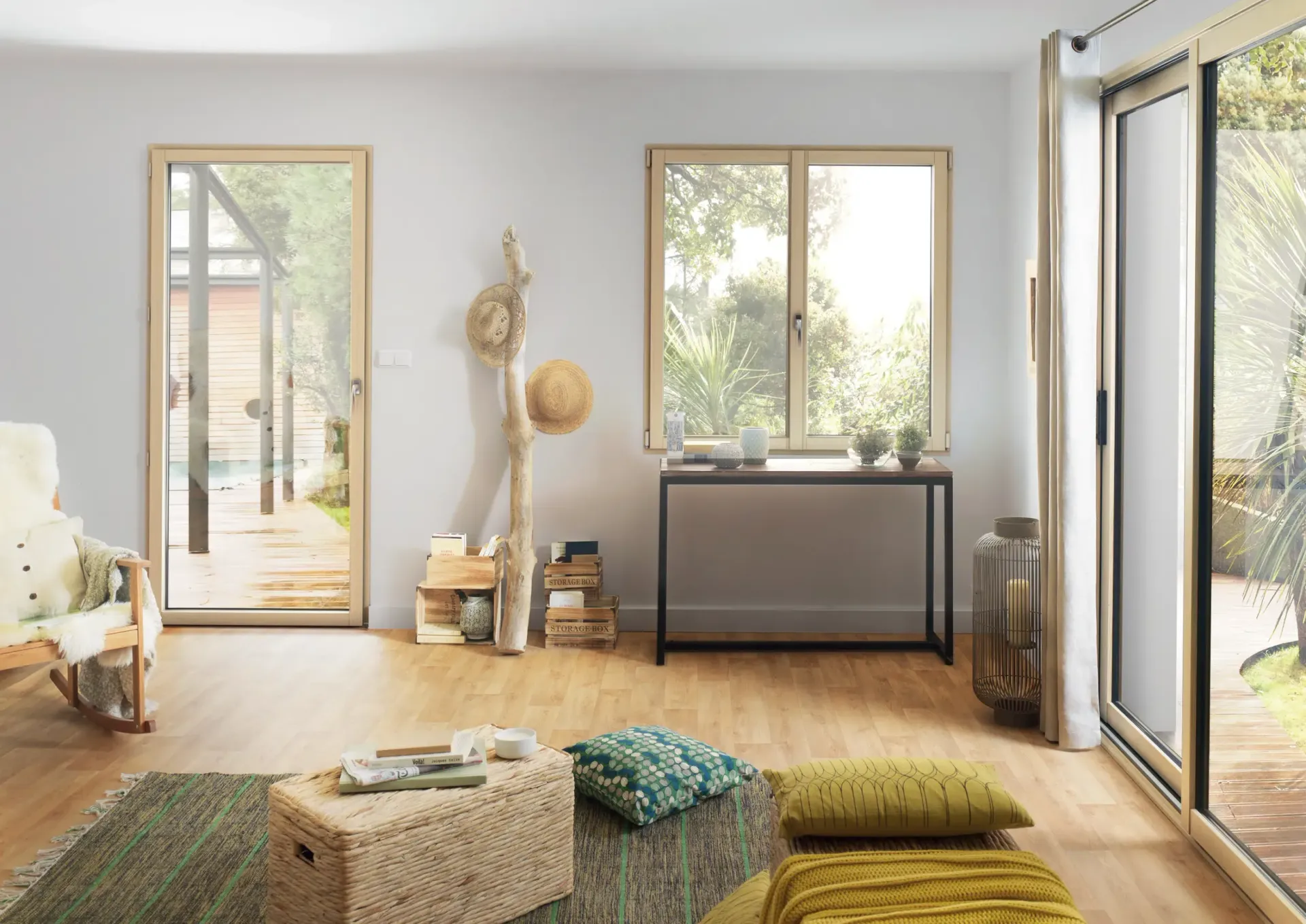 Salon lumineux avec parquet, fenêtres et vue sur l'extérieur ; meublé d'un tapis, de coussins et d'une console.