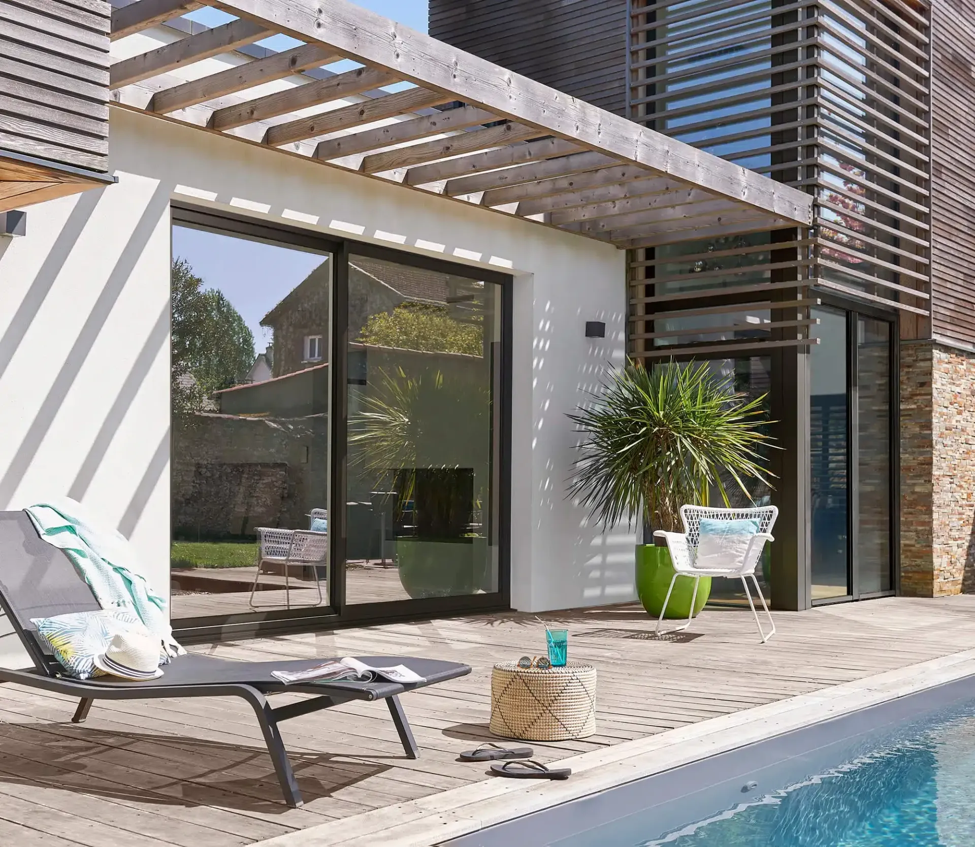 Terrasse avec piscine, transat, chaises et portes coulissantes en verre. Pergola en bois et façade de maison moderne.