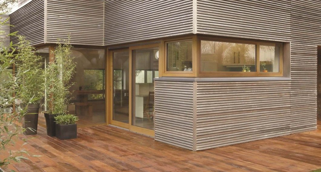 Extérieur de maison moderne en bois avec bardage à claire-voie, terrasse en bois, grandes fenêtres et plantes en pot.