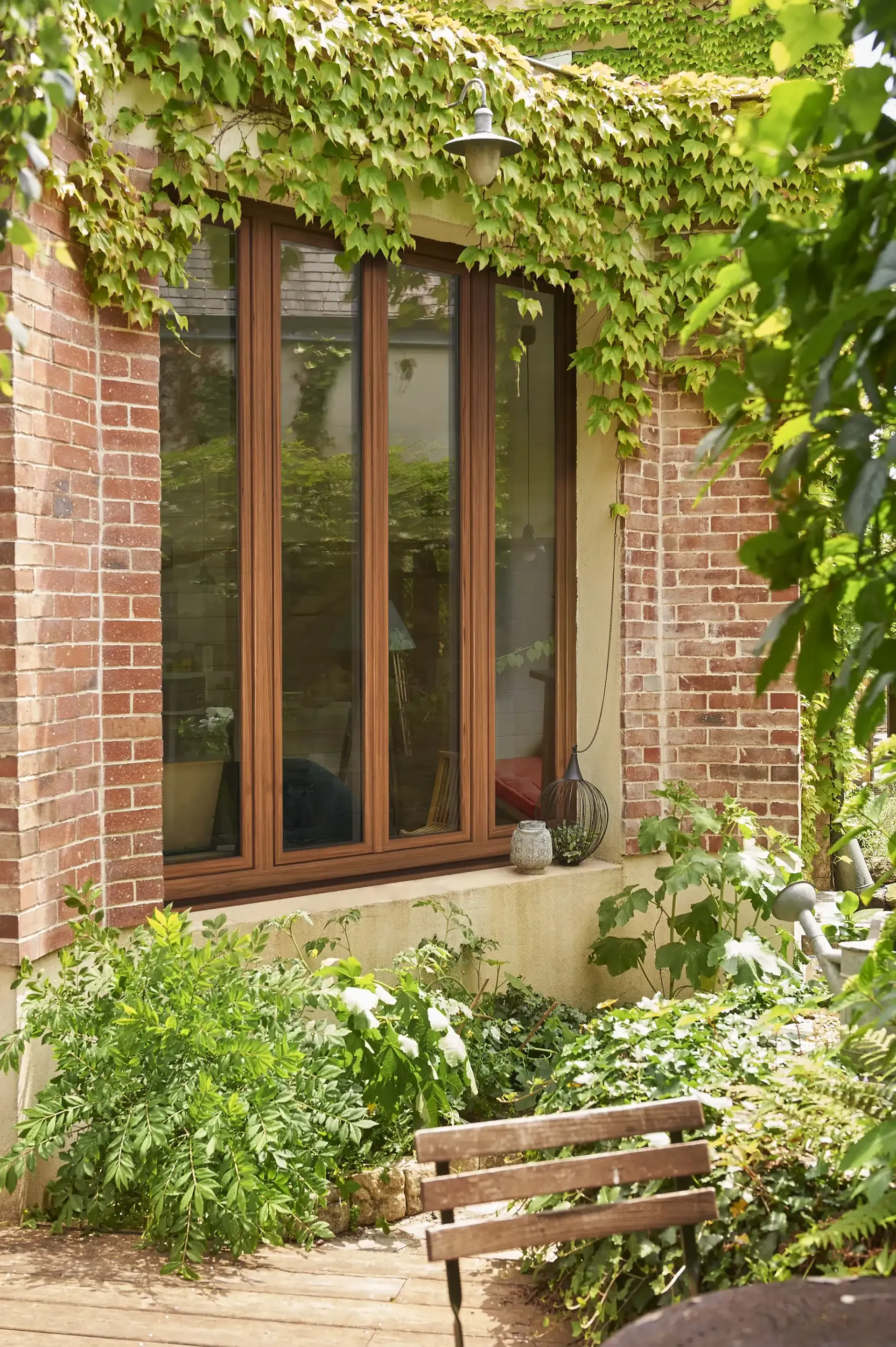 Fenêtre encadrée de briques, recouverte de vigne verte, donnant sur un petit jardin avec un banc.