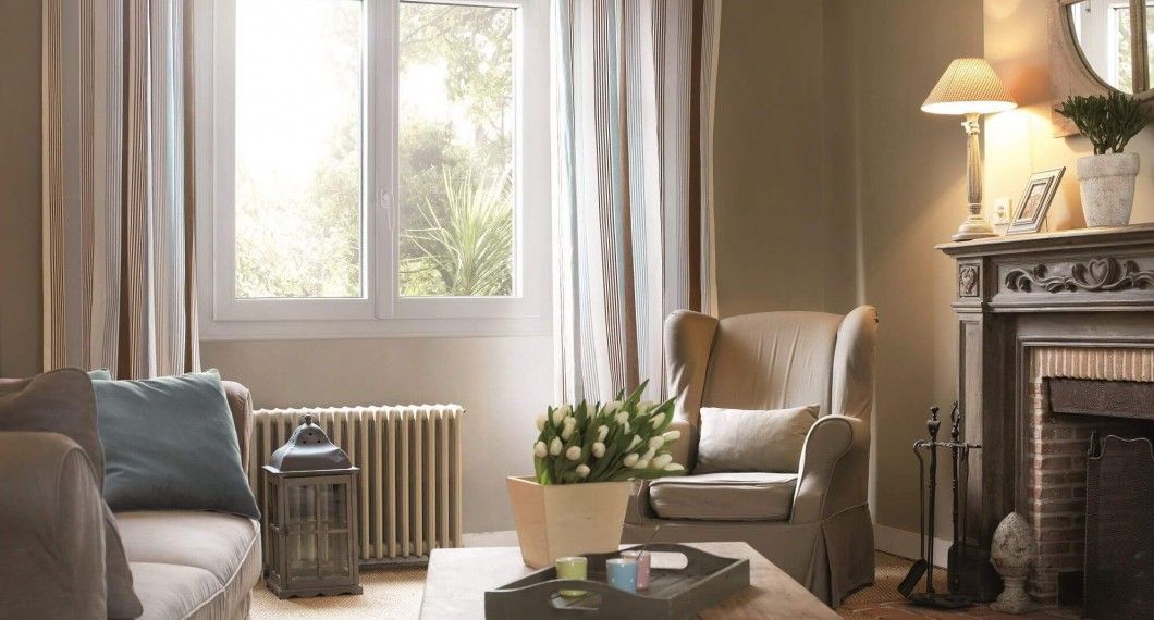 Salon avec fenêtre, cheminée et canapé. Murs beiges, rideaux aux tons neutres, fleurs sur une table.