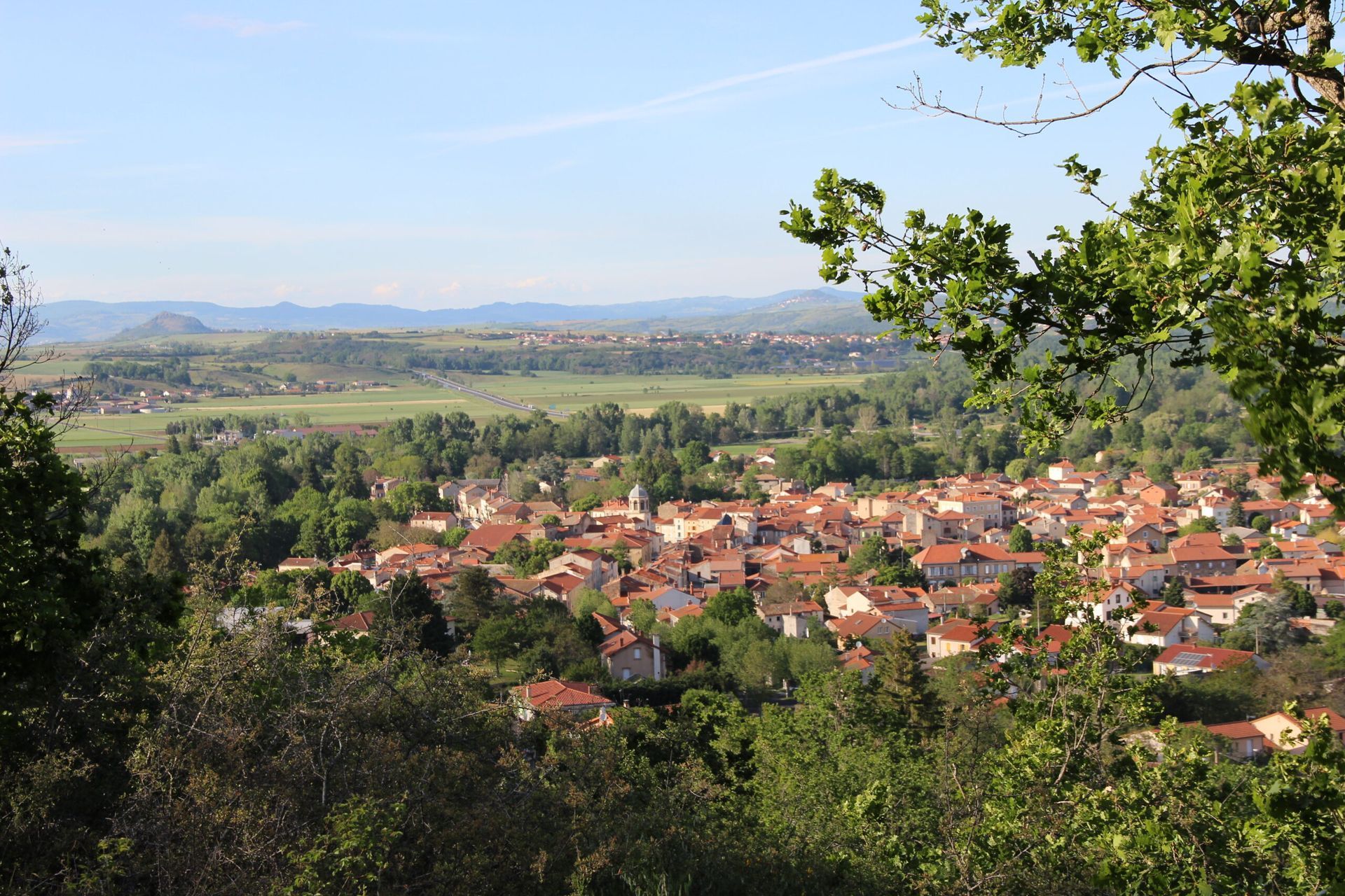 Vue d'un village européen aux toits de tuiles rouges, niché dans une vallée, encadré d'arbres et de collines.