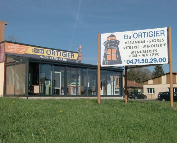 Immeuble avec enseigne Éts Ortigier, proposant vérandas, boutiques et autres commodités. Ciel bleu, pelouse verte.