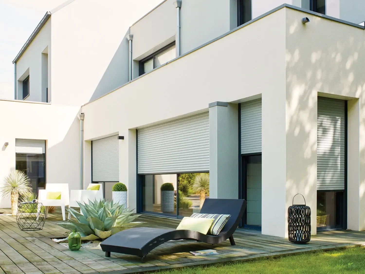 Extérieur d'une maison moderne avec volets roulants blancs partiellement fermés, une chaise longue et une terrasse.
