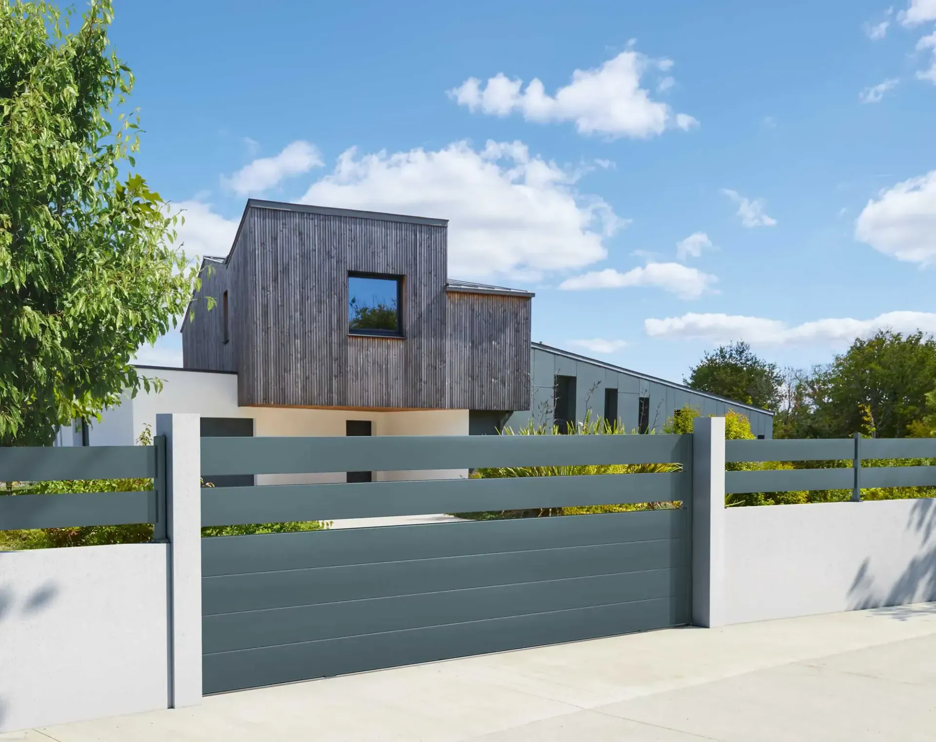 Clôture à lattes horizontales grises avec des poteaux blancs devant une maison moderne revêtue de bois, sous un ciel bleu.