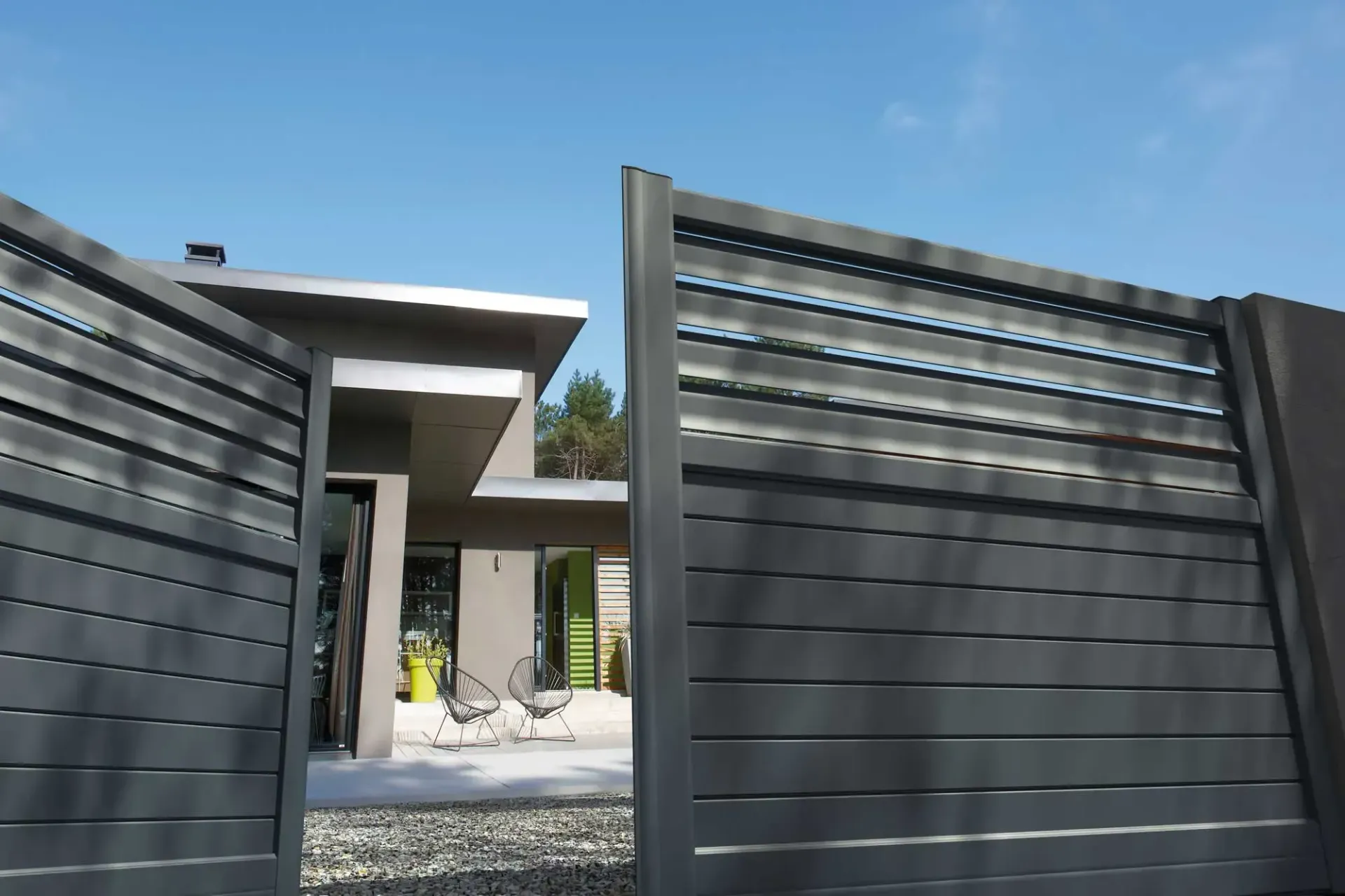 Un portail gris à lattes est partiellement ouvert, dévoilant une maison moderne sur fond de ciel bleu.