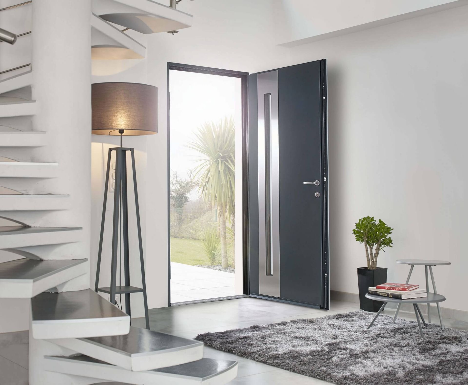 Entrée moderne avec porte gris foncé, ouverte sur l'extérieur. Escalier, lampe, tapis et plante.