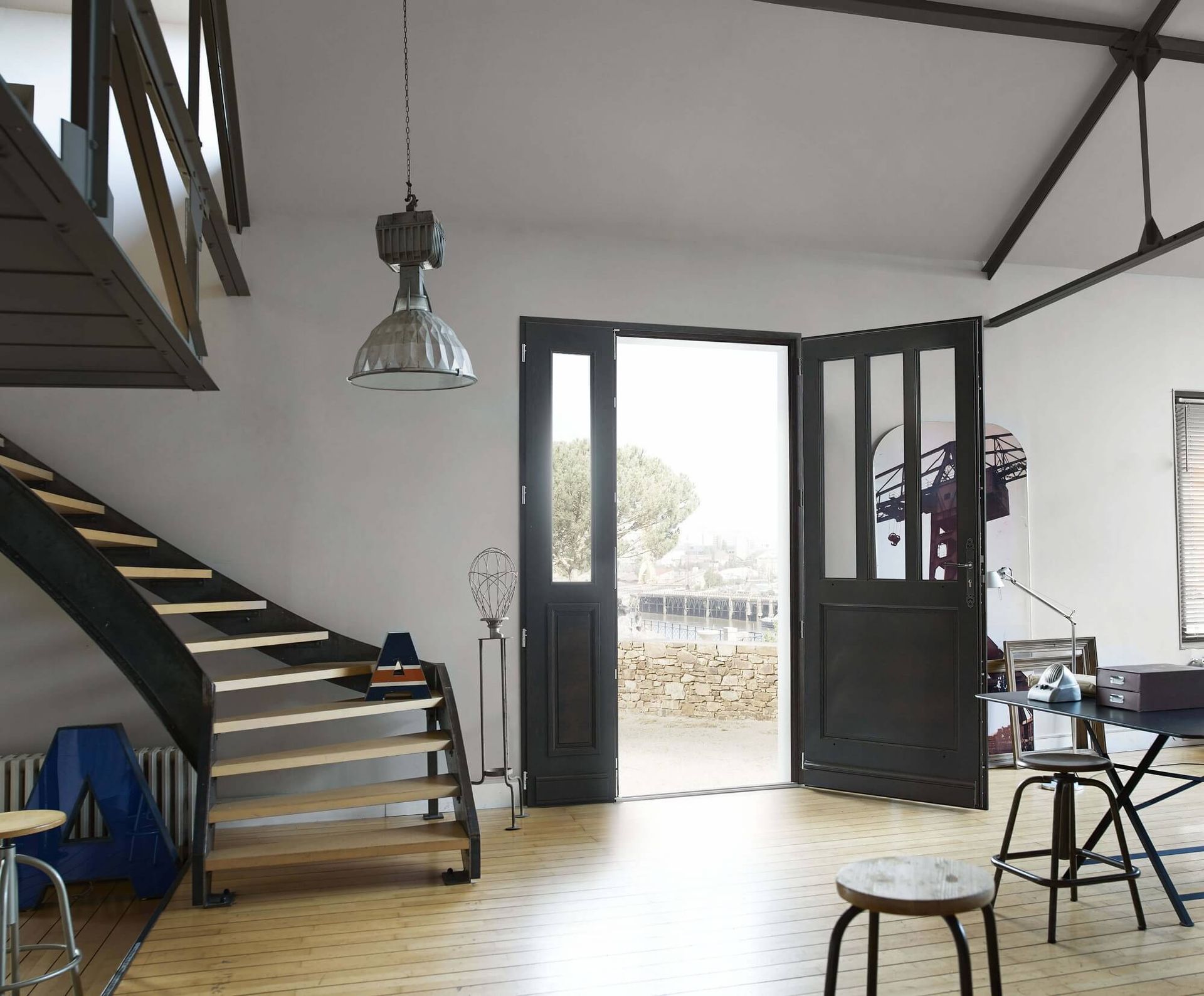 Vue intérieure d'un loft de style industriel avec un escalier, des portes ouvertes sur un espace extérieur et un luminaire suspendu.