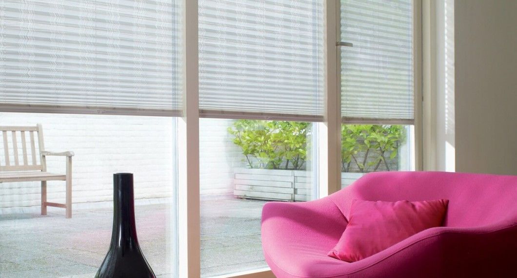 Salon avec stores clairs fermés, vue sur terrasse extérieure meublée de meubles et de plantes en pot, fauteuil rose.