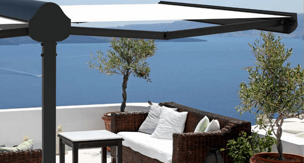 Terrasse avec auvent blanc, canapé en osier, table et arbres en pot donnant sur une mer bleue.