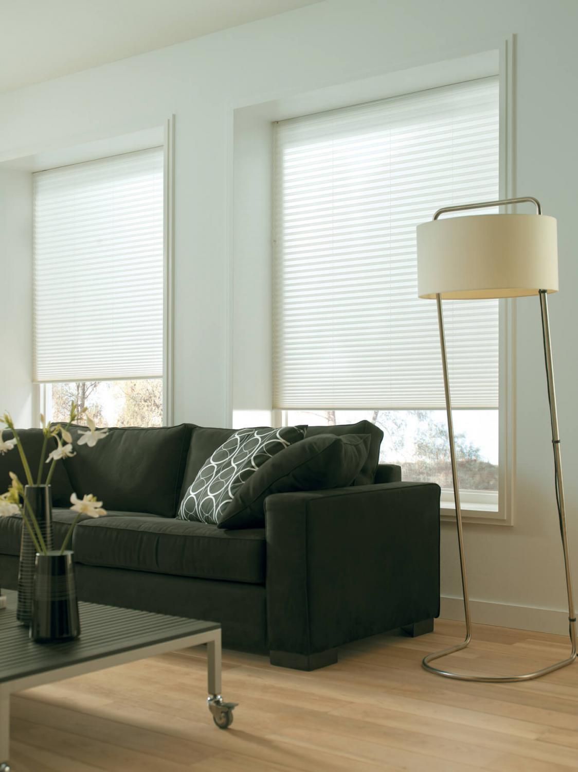 Salon avec un canapé noir, des stores cellulaires clairs et un lampadaire argenté à abat-jour beige.