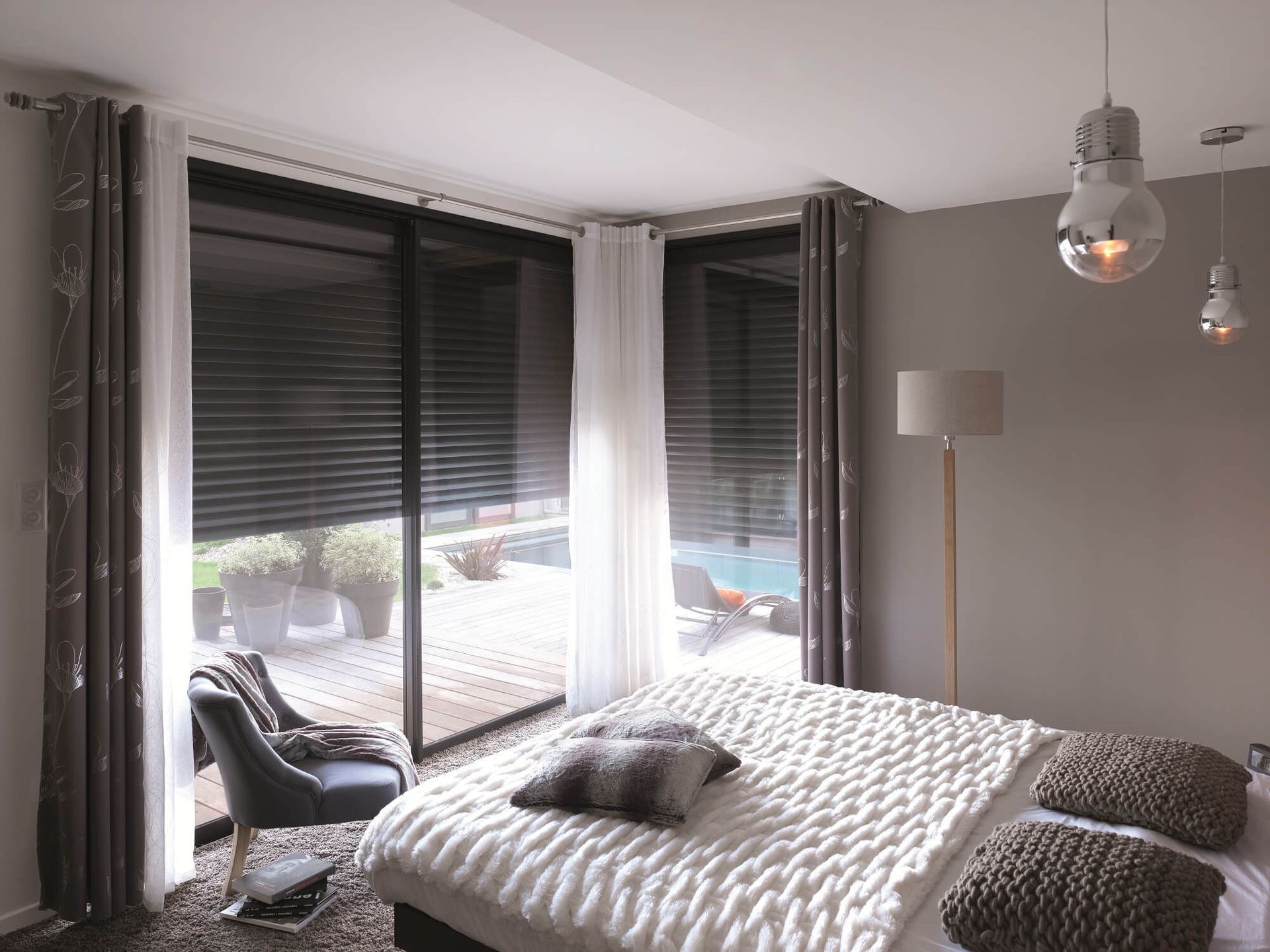 Chambre avec lit, coussins à motifs, voilages, stores et vue sur une terrasse extérieure.