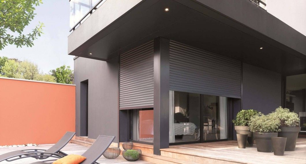 Extérieur de maison moderne avec volets roulants gris partiellement ouverts au-dessus d'une grande fenêtre, patio avec chaises longues.