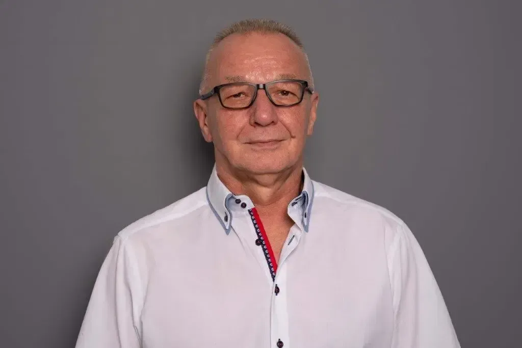 Ein Mann mit Brille und weißem Hemd steht vor einem grauen Hintergrund.